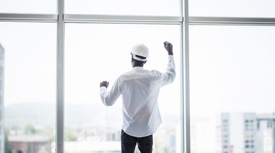 Young afro american man in protective hemlet standing in front of panoramic windows with reaised hands of victory and sucess